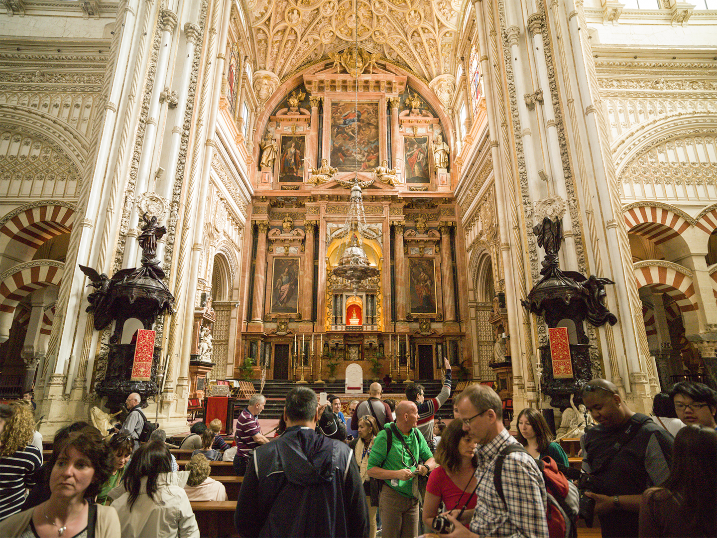 Interior de una catedral monumental con una nave central de gran altura y un altar ricamente decorado al fondo. Numerosos visitantes ocupan los bancos y pasillos, mientras columnas, arcos y detalles ornamentales en tonos claros enmarcan el espacio.