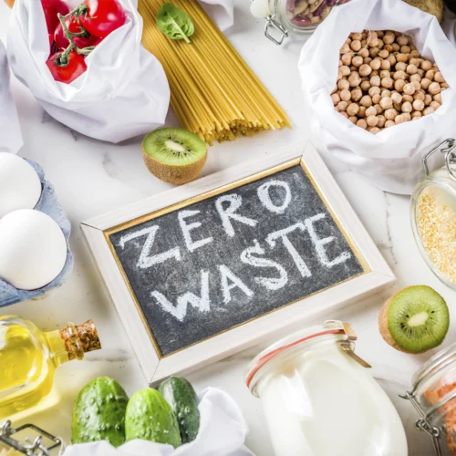 Alimentos a granel y en envases reutilizables rodean una pizarra con el mensaje “ZERO WASTE”. Aparecen huevos, pasta, legumbres, frutas, verduras y tarros de cristal, representando un consumo sostenible y la reducción de residuos.