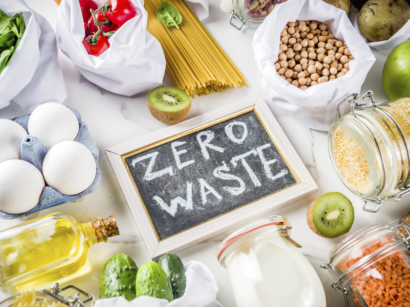 Alimentos a granel y en envases reutilizables rodean una pizarra con el mensaje “ZERO WASTE”. Aparecen huevos, pasta, legumbres, frutas, verduras y tarros de cristal, representando un consumo sostenible y la reducción de residuos.