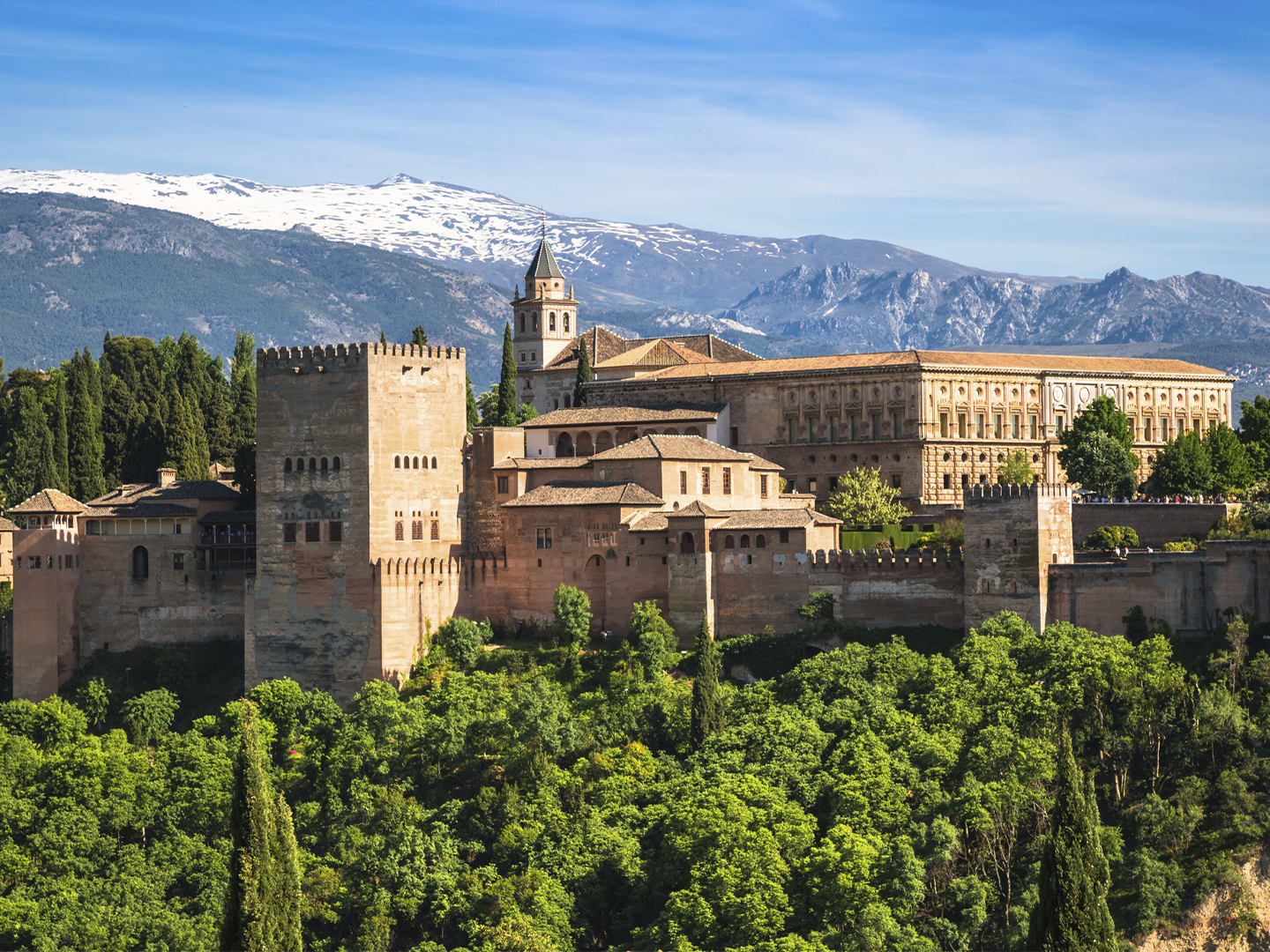 Vista panorámica de un conjunto monumental amurallado rodeado de vegetación, con montañas nevadas al fondo. Las torres y edificios históricos destacan sobre el paisaje bajo un cielo despejado.