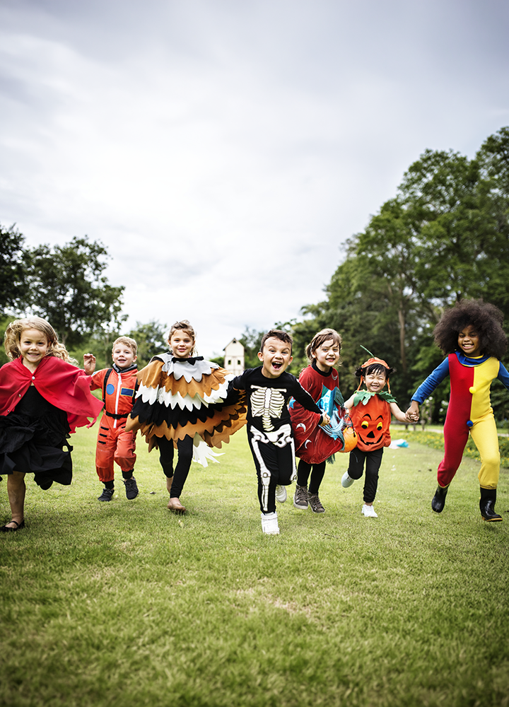 Grupo de niños y niñas corriendo por un campo de césped al aire libre con disfraces de Halloween y fantasía. Los disfraces incluyen una capa roja, un traje de astronauta, un búho con alas, un esqueleto, una calabaza y un traje multicolor. Al fondo se aprecian árboles y un pequeño edificio, bajo un cielo nublado.