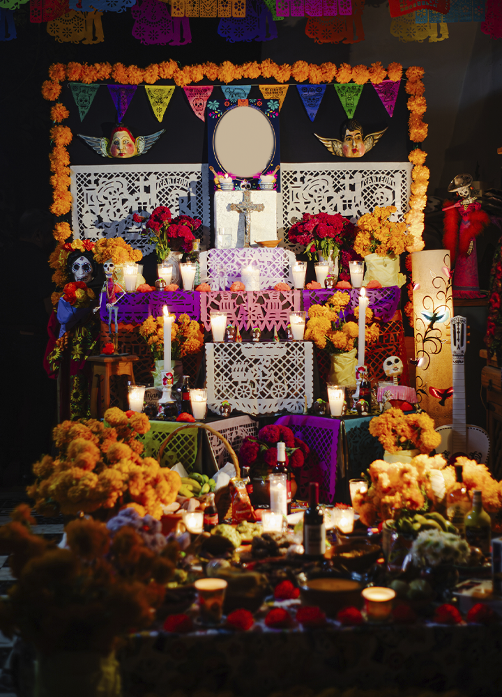 Altar tradicional del Día de Muertos decorado con flores naranjas, velas encendidas, calaveras ornamentales y papel picado de colores. La composición incluye varios niveles con cruces, fotografías enmarcadas, ofrendas de comida y bebida, figuras decorativas y abundantes arreglos florales. La iluminación cálida de las velas contrasta con el fondo oscuro, resaltando los detalles y colores de esta celebración conmemorativa mexicana.