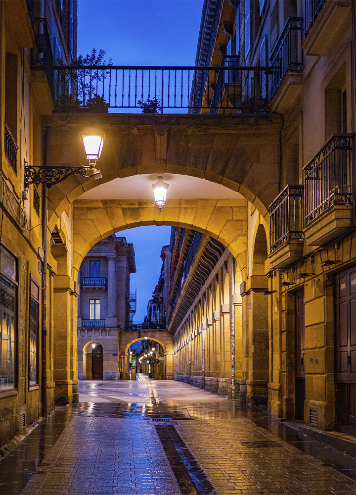 Calle empedrada con soportales y arcos de piedra iluminados por farolas, reflejados sobre el suelo mojado al atardecer.