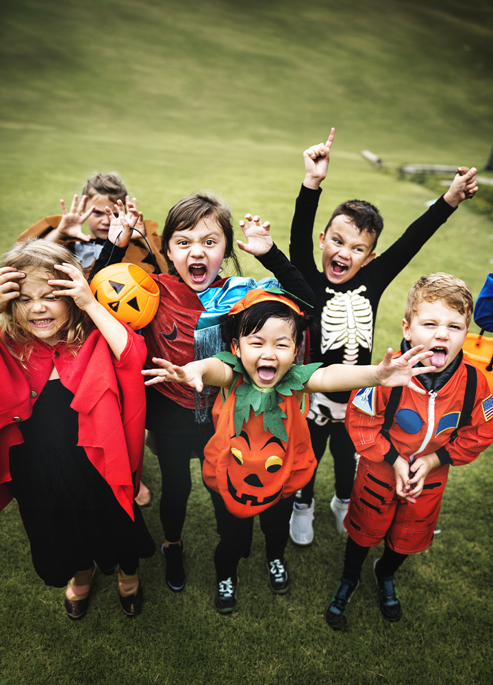 Grupo de niños y niñas disfrazados para Halloween posando sobre el césped al aire libre. Los disfraces incluyen una capa roja, una calabaza, un esqueleto, un traje espacial y una capa de superhéroe. Algunas personas sostienen calabazas decorativas mientras extienden los brazos hacia la cámara en una escena festiva y colorida.
