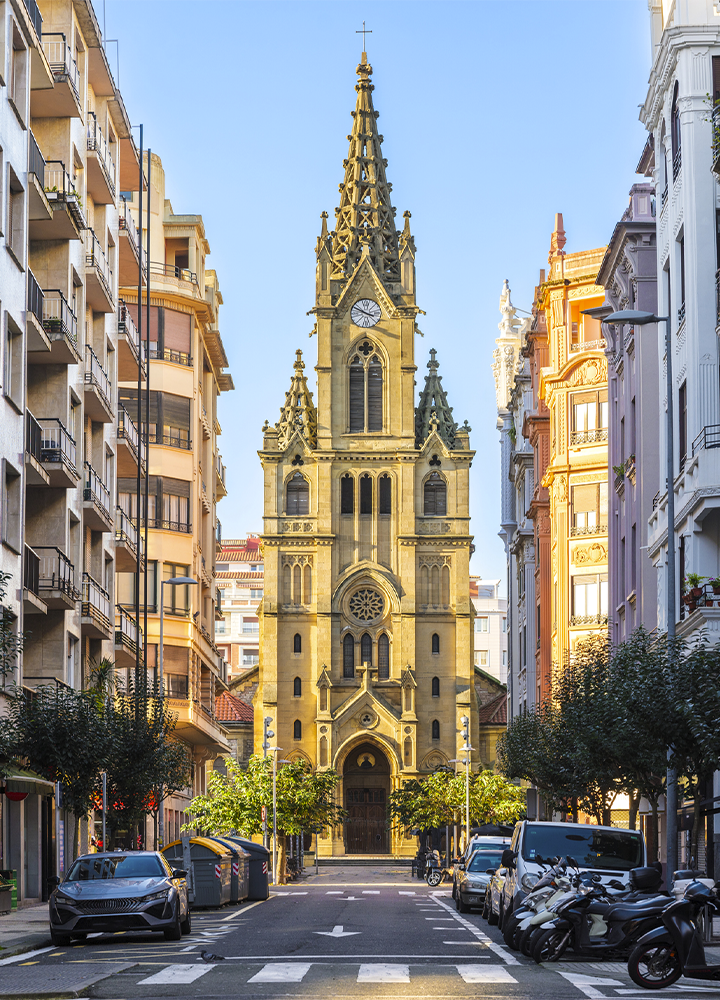 Iglesia de estilo neogótico con una alta aguja central y reloj, situada al final de una calle urbana flanqueada por edificios residenciales y vehículos aparcados.