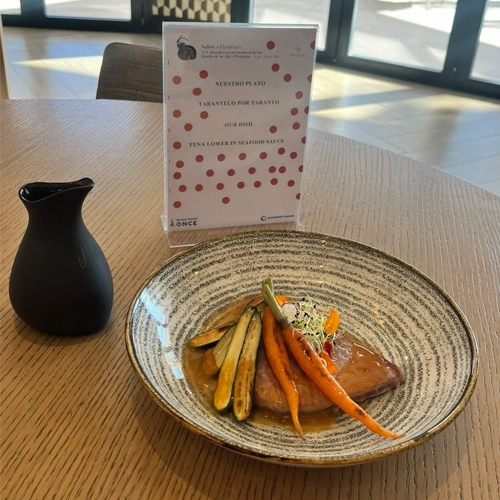 Plato con verduras asadas y salsa servido sobre una mesa, acompañado de una pequeña jarra negra. Al fondo, un cartel identifica la propuesta gastronómica y destaca el uso de sabores y productos de temporada.