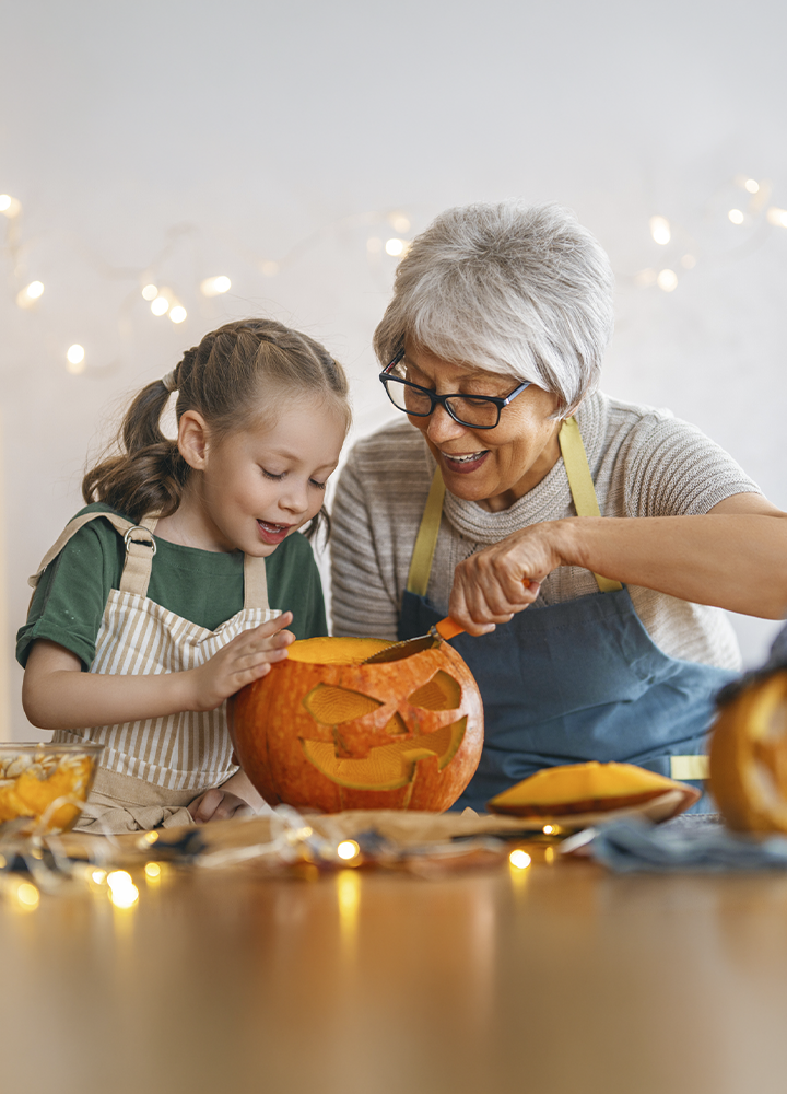 Dos personas con delantales tallan una calabaza de Halloween sobre una mesa decorada con luces cálidas. Una de las personas utiliza una herramienta de corte mientras la otra observa los detalles de la calabaza ya vaciada. Alrededor se aprecian trozos de pulpa, utensilios y otra calabaza parcialmente visible, en un ambiente doméstico preparado para la celebración de Halloween.