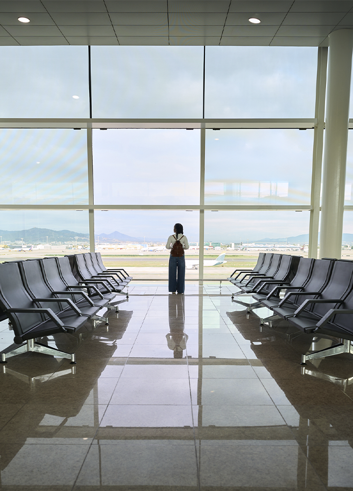Sala de espera de un aeropuerto con filas de asientos vacíos a ambos lados de un amplio pasillo. Al fondo, una persona observa el exterior a través de un gran ventanal que ofrece vistas a la pista de aterrizaje y a varios aviones. El suelo pulido refleja la luz natural y la estructura simétrica del espacio, creando una sensación de amplitud y calma. Proporcione sus comentarios sobre BizChat Hazlo más cortoRevisa el alt