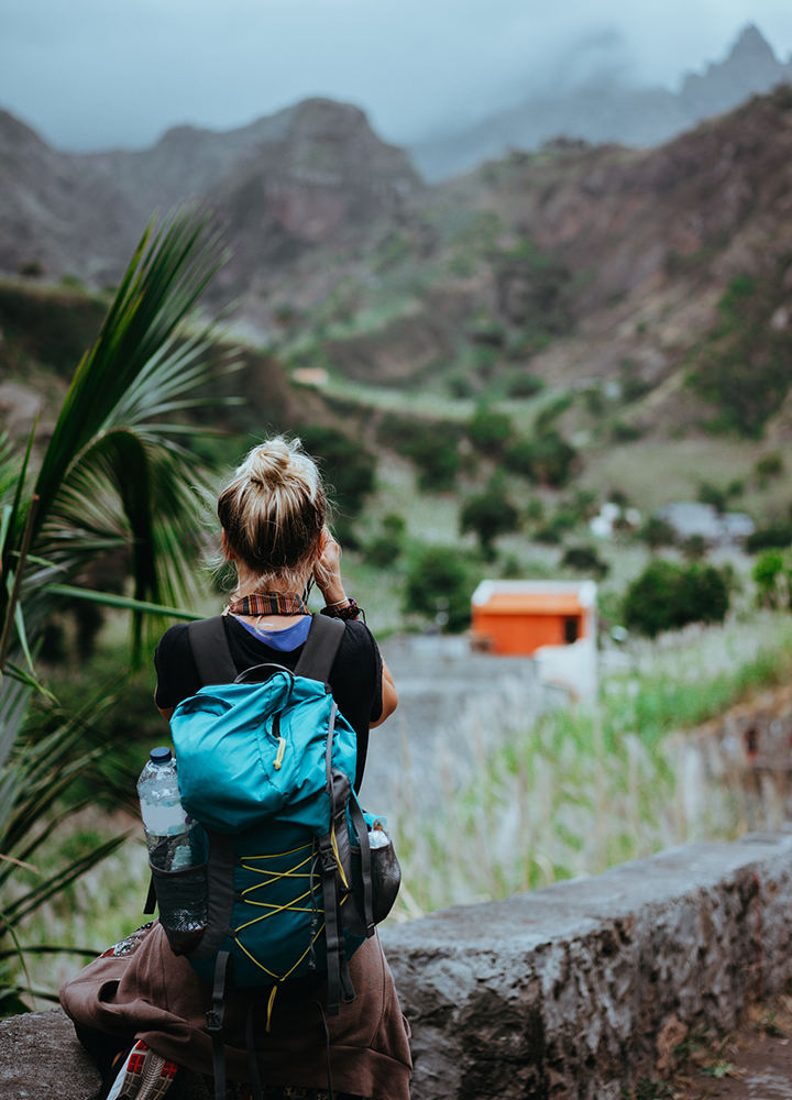 Persona con mochila contempla un paisaje montañoso desde un mirador, con vegetación y construcciones dispersas en el valle al fondo.