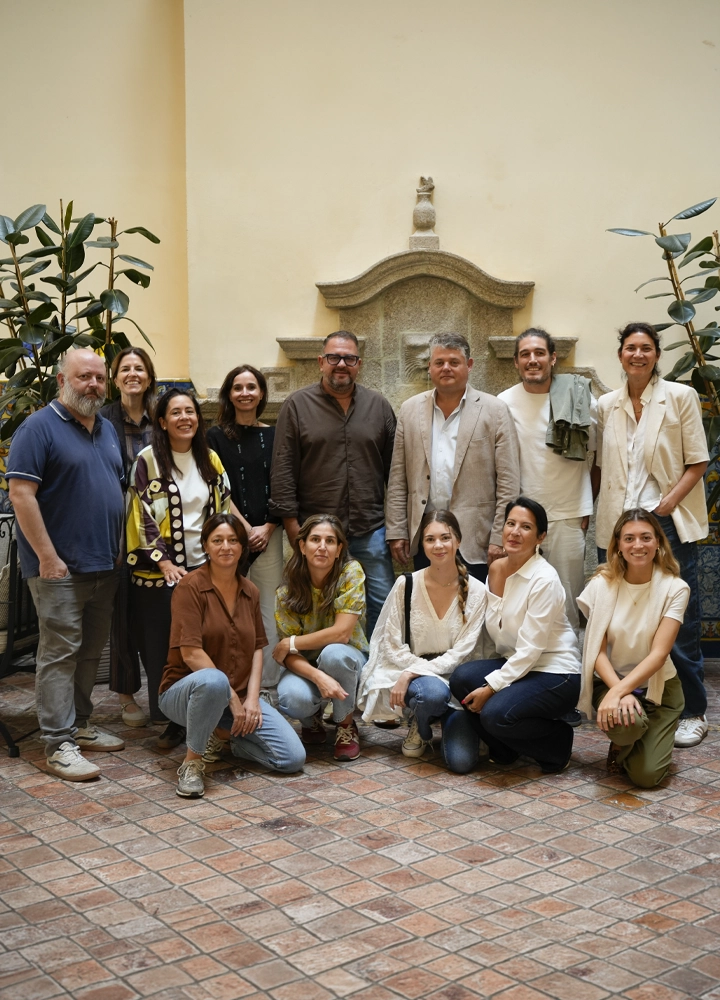 Grupo de trece personas posando en un patio interior con suelo de baldosas, plantas ornamentales y una fuente de piedra en la pared. Varias personas aparecen de pie y otras agachadas en primera fila.