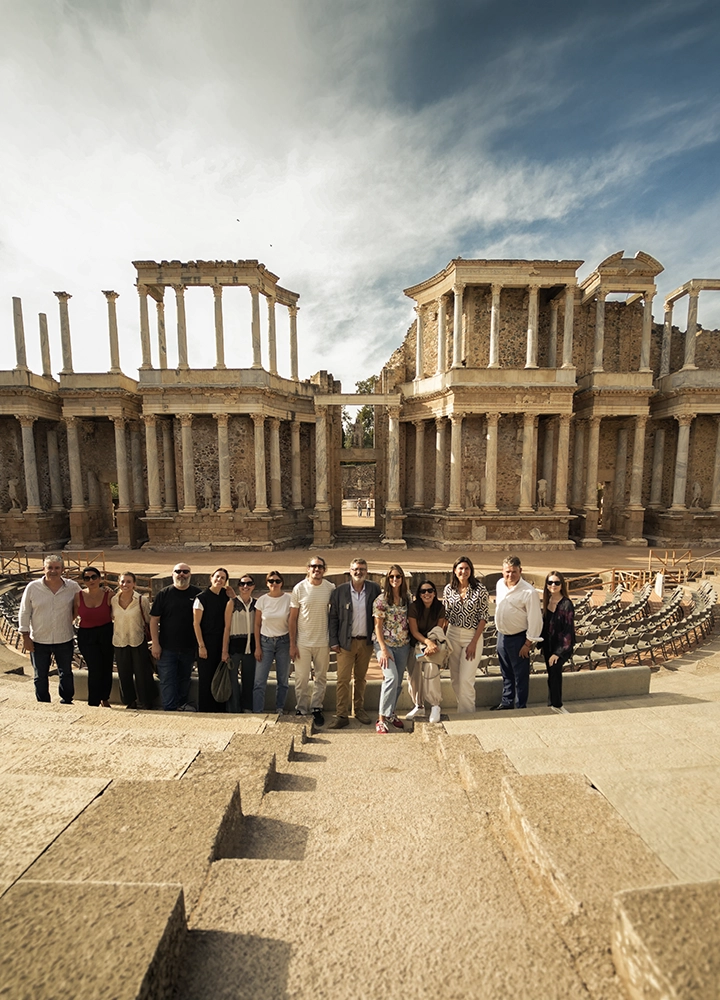 Grupo de personas posando en las gradas de un teatro romano frente a un escenario monumental con columnas y ruinas históricas. El conjunto aparece durante una visita cultural al aire libre.