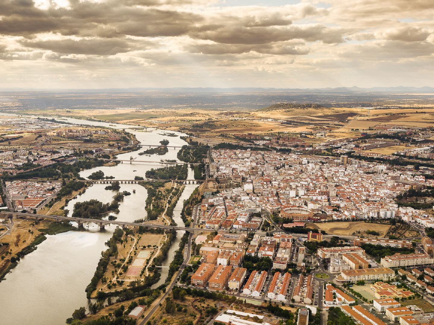 Vista aérea de una ciudad atravesada por un río con varios puentes, rodeada de zonas urbanas y campos agrícolas. Nubes densas cubren el cielo sobre el paisaje.