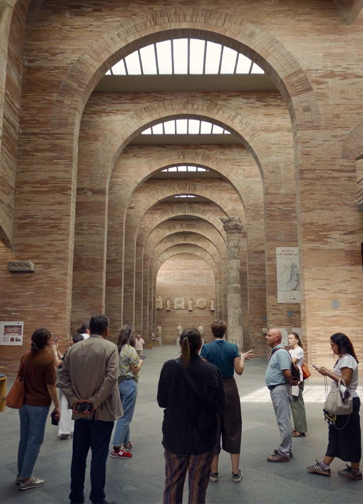 Grupo de personas visitando el interior de un museo o espacio histórico de grandes dimensiones, con altos arcos de ladrillo, columnas y esculturas al fondo. La guía se dirige al grupo durante el recorrido.