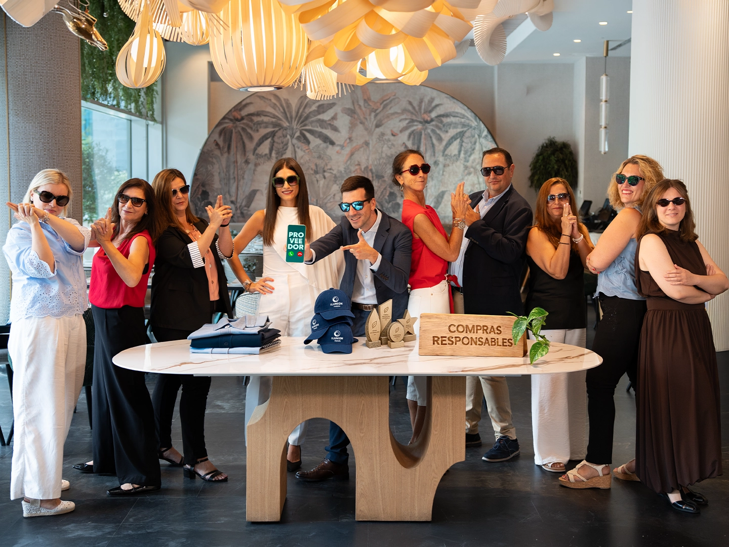 Grupo de diez personas posando alrededor de una mesa con artículos promocionales, trofeos y un cartel con el texto “Compras Responsables”. Todas llevan gafas de sol y adoptan una pose lúdica en el vestíbulo de un edificio moderno con lámparas decorativas colgantes.