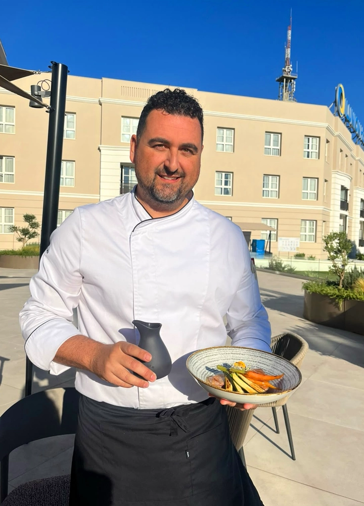 Persona con chaqueta de chef sostiene un plato con verduras y una pequeña jarra en una terraza exterior, con un edificio hotelero al fondo bajo un cielo despejado.
