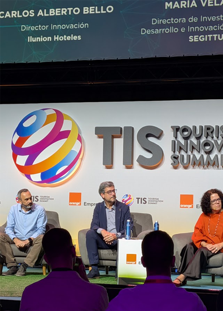 Participantes de una mesa redonda intervienen en el Tourism Innovation Summit (TIS), sentados en un escenario frente al logotipo del evento y una pantalla con información de los ponentes.