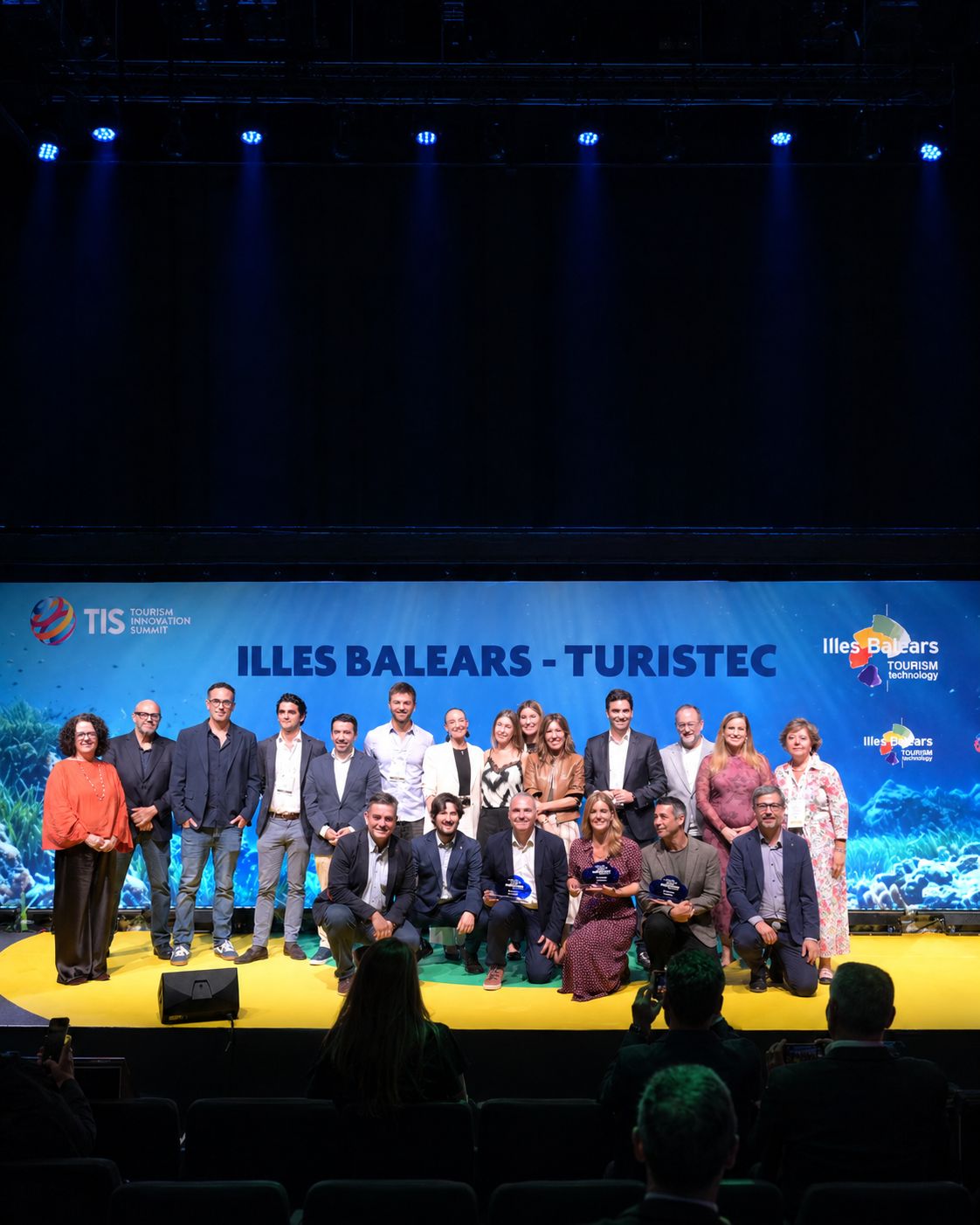 Grupo de personas posa sobre un escenario durante una entrega de premios, con varios galardonados en primera fila y un gran panel azul al fondo con el texto «Illes Balears - Turistec».