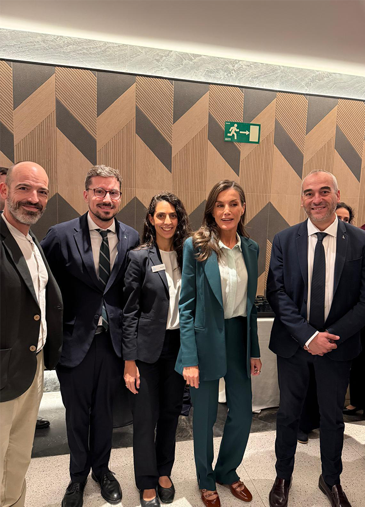 Cinco personas posan juntas en un espacio interior durante un evento corporativo. Visten trajes y ropa formal frente a una pared con diseño geométrico de madera y una señal de salida de emergencia visible en la parte superior.