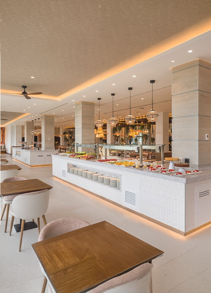 Restaurante de hotel con diseño contemporáneo y una amplia zona de bufé central. El espacio cuenta con mostradores blancos repletos de alimentos, mesas de madera, sillas tapizadas en tonos claros y lámparas colgantes decorativas. La iluminación cálida, las columnas revestidas en piedra y la presencia de vegetación crean un ambiente luminoso y acogedor.