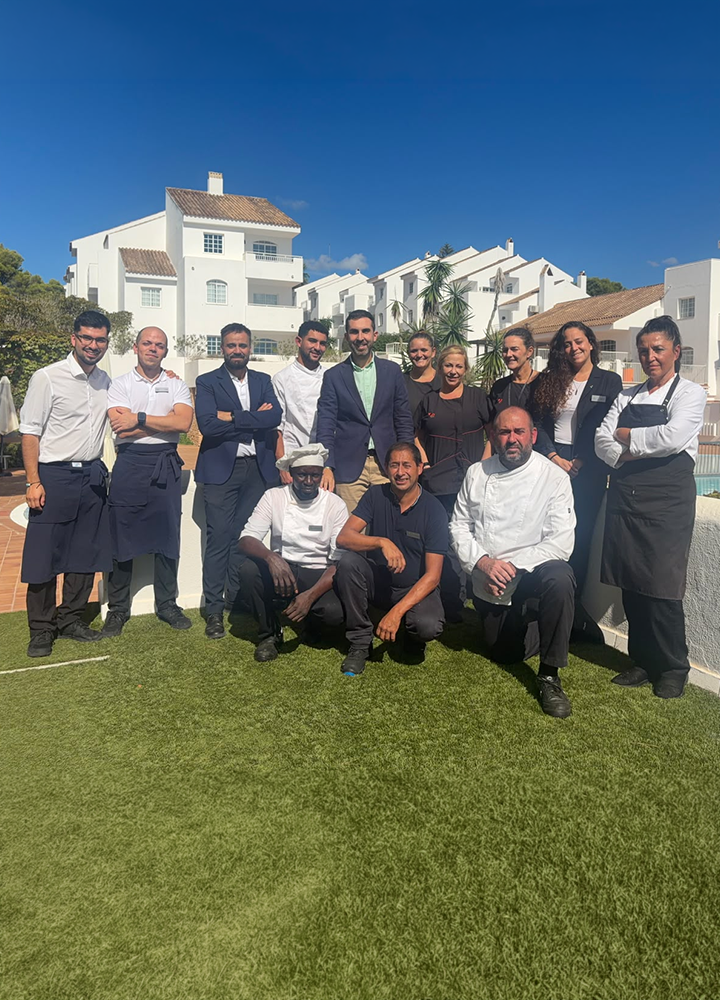 Grupo de once personas posando sobre césped en un complejo de edificios blancos, bajo un cielo despejado y soleado.
