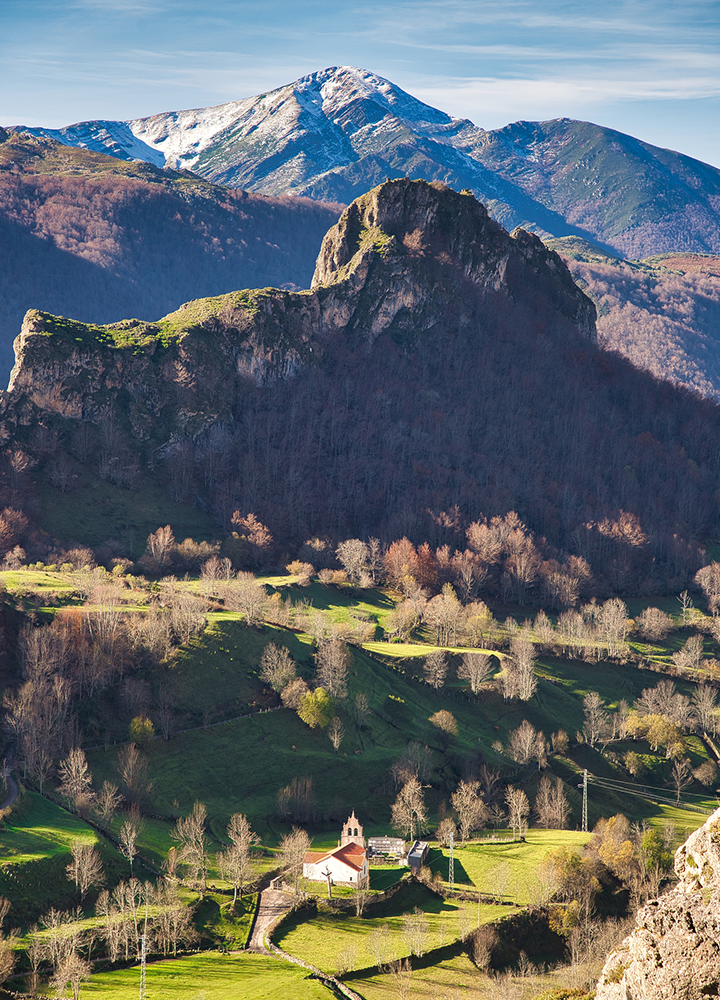 Paisaje de montaña con un pequeño edificio rodeado de praderas y árboles en primer plano. Al fondo, una formación rocosa y cumbres elevadas dominan el horizonte bajo un cielo despejado.