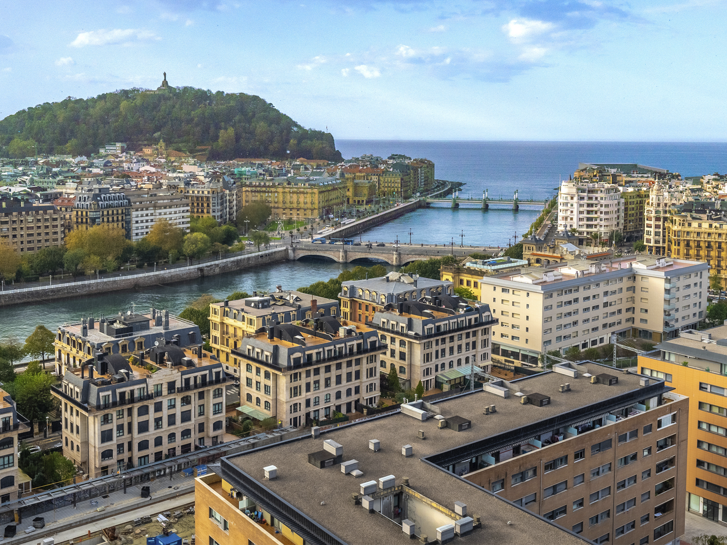 Vista panorámica de San Sebastián desde un edificio elevado, con el río Urumea, varios puentes, edificios urbanos y el mar al fondo. A la izquierda destaca el monte Urgull coronado por una estatua.