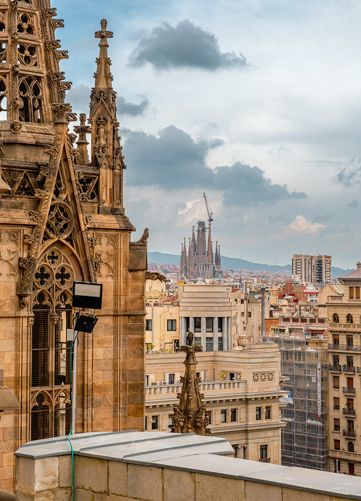 Vista urbana desde una terraza o mirador, con detalles ornamentales de una fachada histórica en primer plano y la Sagrada Familia al fondo, elevándose sobre los edificios de la ciudad bajo un cielo nublado.