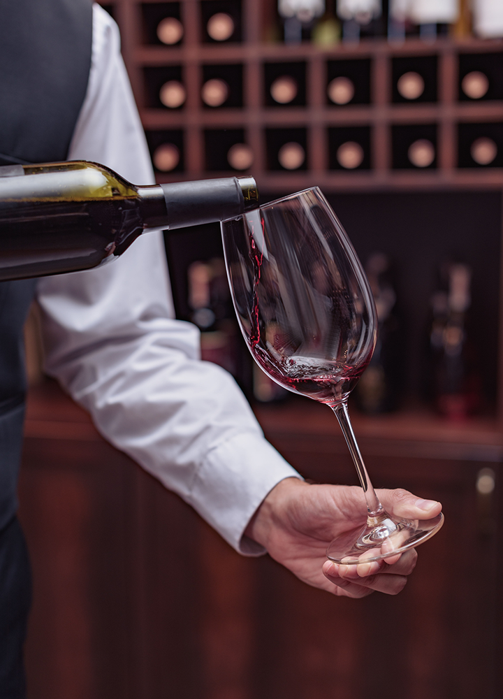 Servicio de vino tinto en una copa, con una botella inclinada y una vinoteca desenfocada al fondo.