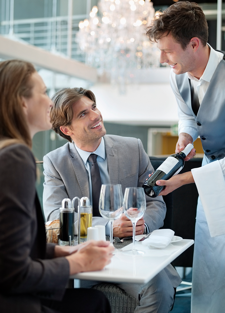 Personal de sala presenta una botella de vino a dos comensales sentados en la mesa de un restaurante, con copas preparadas para el servicio.
