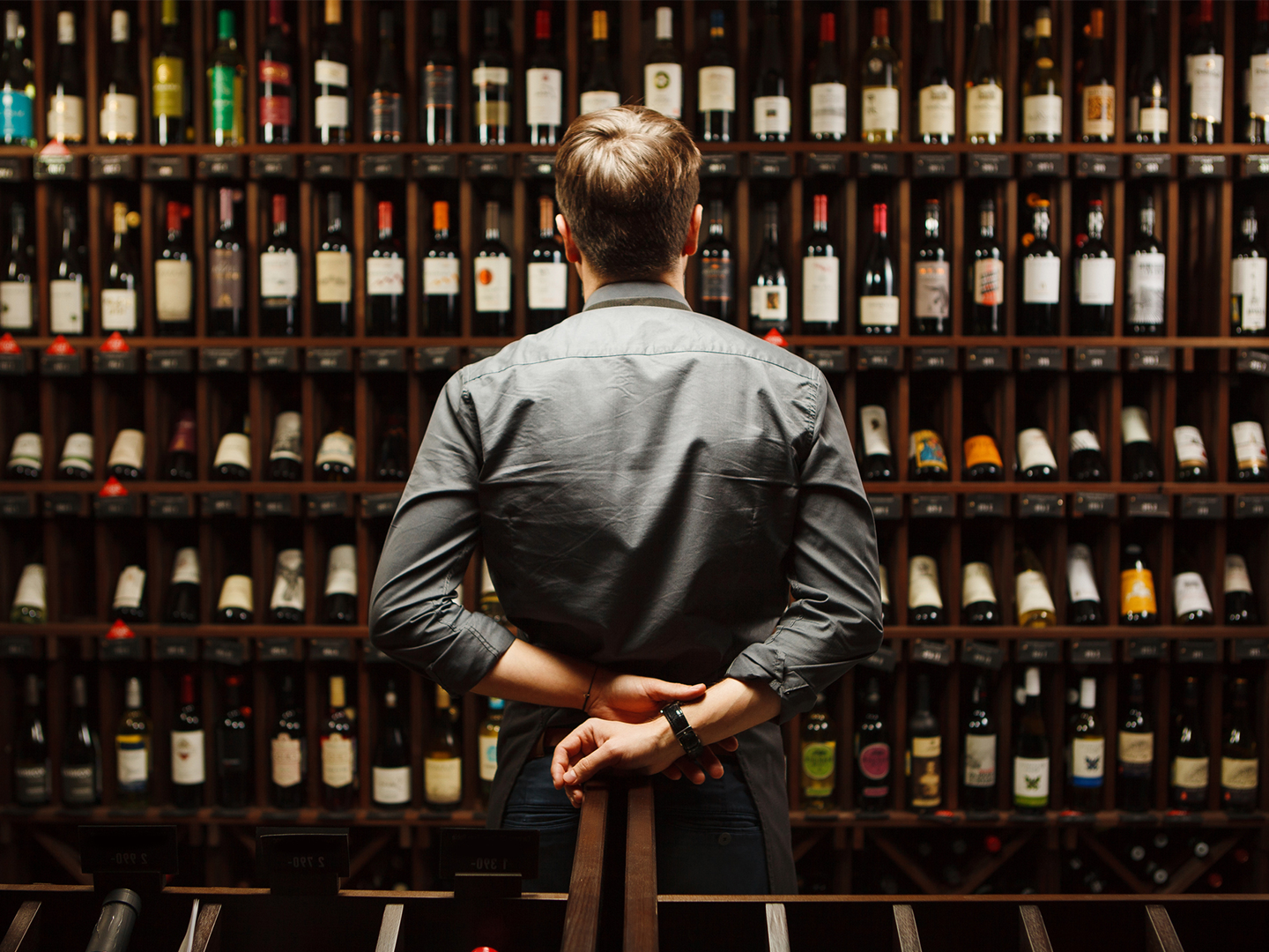 Persona de espaldas observa una pared repleta de botellas de vino organizadas en estanterías de madera dentro de una vinoteca.